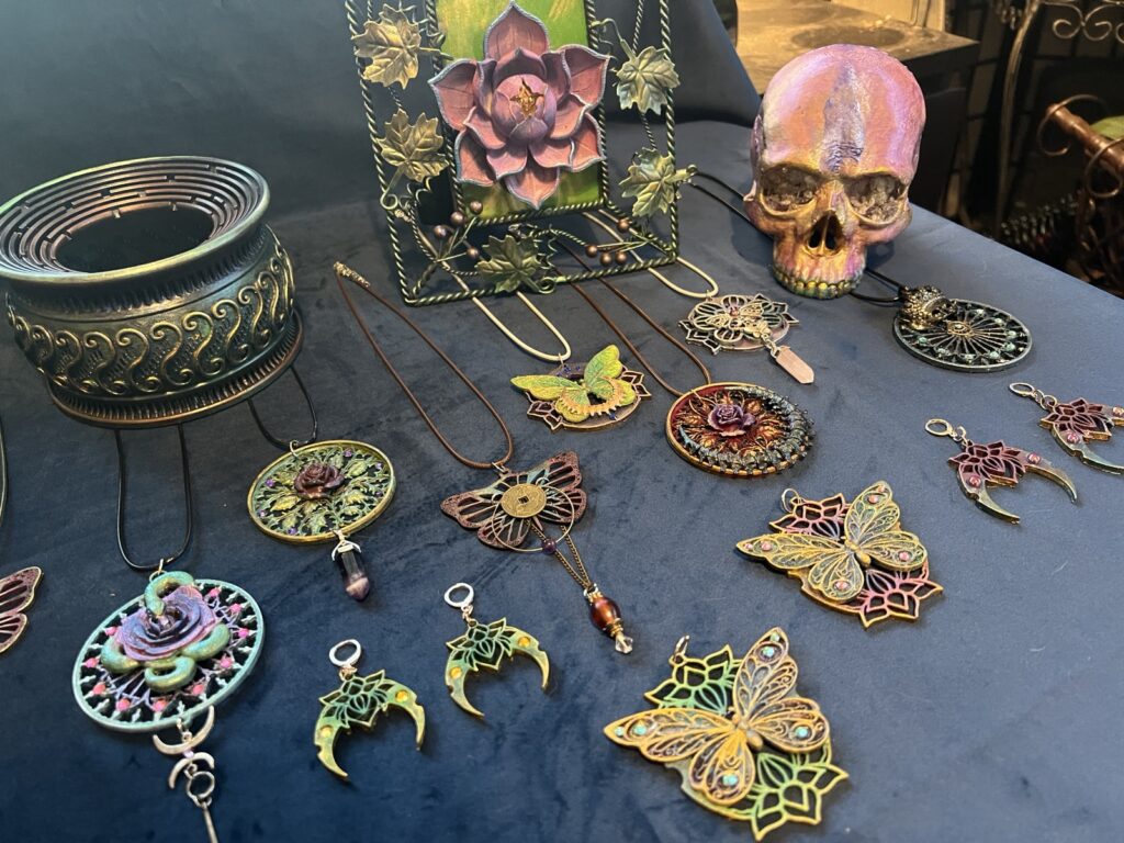 Handmade pendants, earrings, and painted objects arranged on dark blue velvet, including butterfly pendants, crescent moon earrings, an ornamented vessel, and a painted skull.