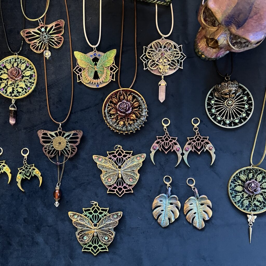 Layered pendants and earrings on dark velvet, including filigree butterflies, rose medallions, crescent moons, and monstera leaf earrings in painted metallic finishes.