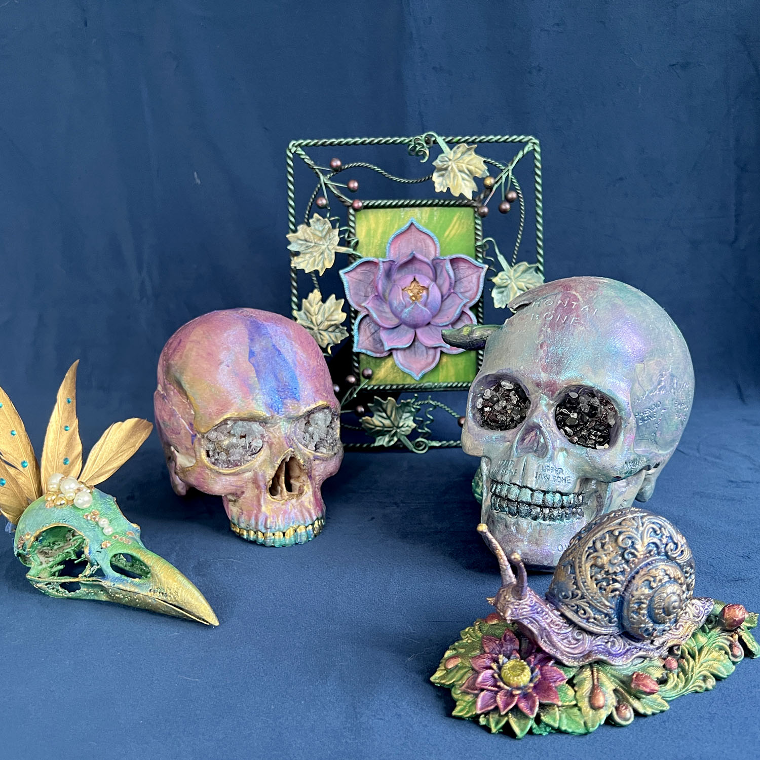 Painted skulls and sculpted objects on dark blue velvet: an iridescent skull with crystal-filled eye sockets and anatomical labels, a purple and gold skull with crystal eyes, a green raven skull crowned with gold feathers and pearls, an ornate snail on a bed of painted flowers, and a framed lotus panel behind.