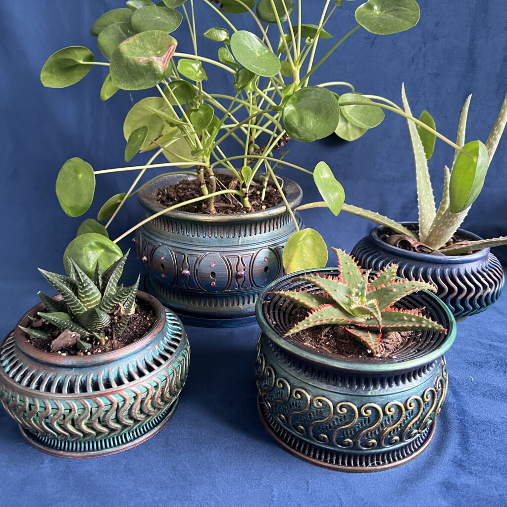 Four ornamental planters with sculpted relief patterns on dark blue velvet, finished in verdigris, copper, gold, and deep blue patina, holding a pilea, an aloe, and two small succulents.